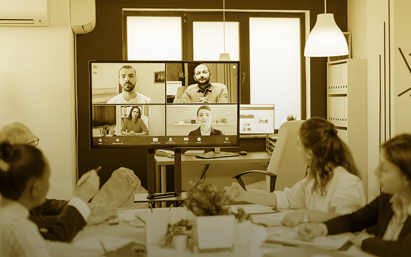 A imagem ilustra uma sala de reuniões com diversas pessoas, sendo algumas de forma presencial e outras em uma vídeo chamada.